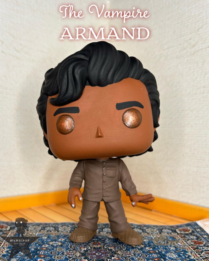 May include: A vinyl collectible figure of The Vampire ARMAND with dark wavy hair, brown skin, and metallic copper eyes. The figure wears a brown button-down shirt and matching trousers. The text The Vampire ARMAND is displayed above the figure.