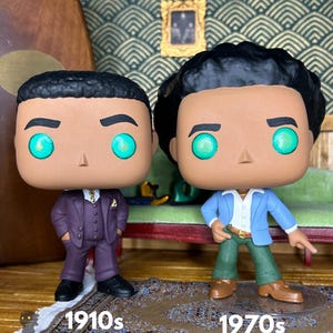 May include: Two vinyl figures with large heads and glowing green eyes. The left figure wears a dark purple suit, while the right figure wears a light blue blazer, white shirt, and green trousers. Text below reads 1910s and 1970s respectively.