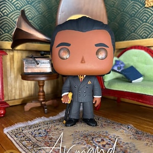 May include: A stylized vinyl collectible figure of a person with dark hair, wearing a charcoal gray pinstriped double-breasted suit, a yellow and red striped tie, and a pocket square. The figure holds a gold-topped black cane in the right hand.