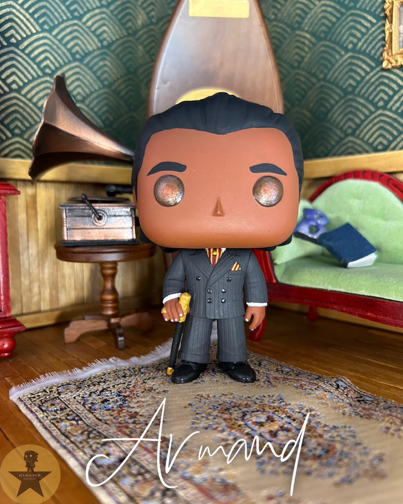 May include: A stylized vinyl collectible figure of a person with dark hair, wearing a charcoal gray pinstriped double-breasted suit, a yellow and red striped tie, and a pocket square. The figure holds a gold-topped black cane in the right hand.