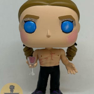 May include: A stylized vinyl figure with light brown hair in two braids, bright blue eyes, and pink cheeks. The figure is shirtless with visible chest markings, wearing black pants and dark shoes, holding a small clear glass with red liquid.
