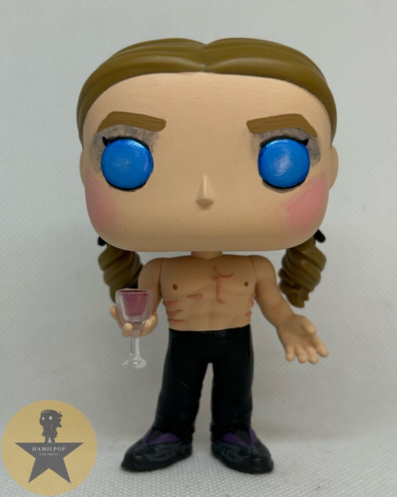 May include: A stylized vinyl figure with light brown hair in two braids, bright blue eyes, and pink cheeks. The figure is shirtless with visible chest markings, wearing black pants and dark shoes, holding a small clear glass with red liquid.