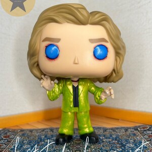 May include: A stylized vinyl figure with blonde hair, bright blue eyes, and a metallic lime green suit over a black shirt. The figure has a small red detail around the neck and is posed with hands raised. The text "Lestat de Lioncourt" is written below.