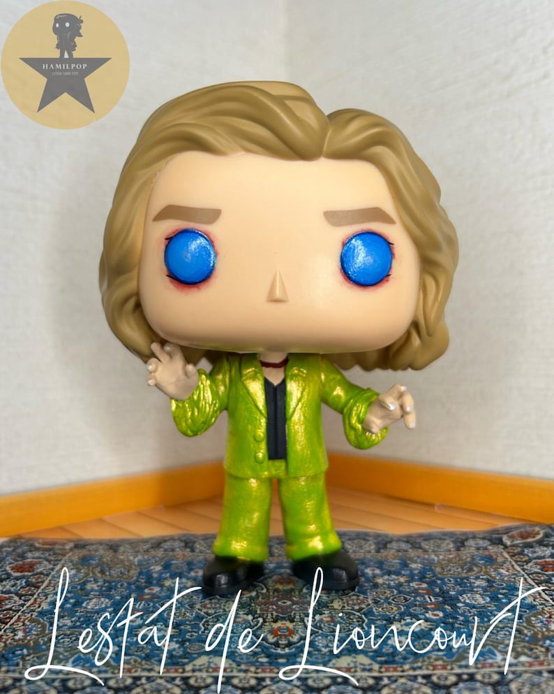 May include: A stylized vinyl figure with blonde hair, bright blue eyes, and a metallic lime green suit over a black shirt. The figure has a small red detail around the neck and is posed with hands raised. The text "Lestat de Lioncourt" is written below.