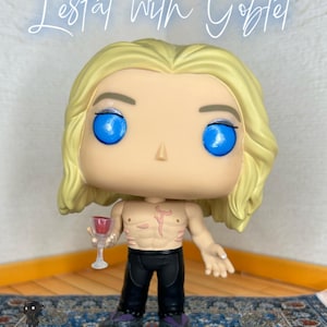 May include: A vinyl collectible figure of a character with long blonde hair, bright blue eyes, and a bare chest featuring a red mark. The figure wears black pants and holds a small clear goblet with red liquid. Text above reads: Lestat with Goblet.