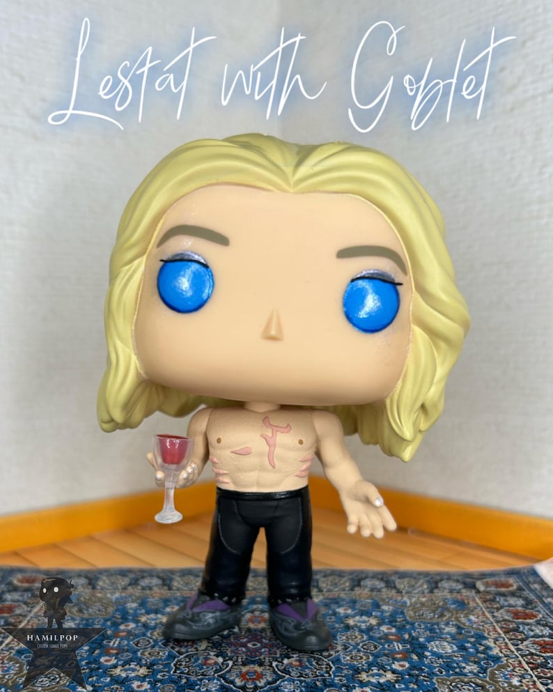 May include: A vinyl collectible figure of a character with long blonde hair, bright blue eyes, and a bare chest featuring a red mark. The figure wears black pants and holds a small clear goblet with red liquid. Text above reads: Lestat with Goblet.