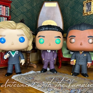 May include: Three stylized vinyl figures representing characters from Interview with the Vampire. The left figure has blonde hair and a grey suit, the center figure has dark hair and a purple suit, and the right figure has dark hair and a grey suit.