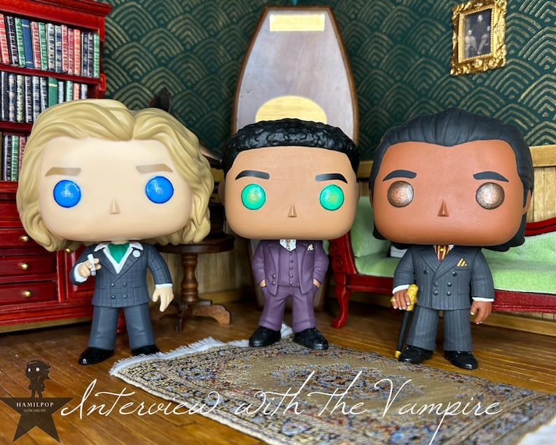 May include: Three stylized vinyl figures representing characters from Interview with the Vampire. The left figure has blonde hair and a grey suit, the center figure has dark hair and a purple suit, and the right figure has dark hair and a grey suit.