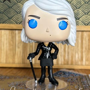 May include: A stylized vinyl collectible figure with white hair, bright blue eyes, and a small beauty mark. The figure wears a black leather-style jacket, a black skirt, and black boots, while holding a black cane with a silver handle.