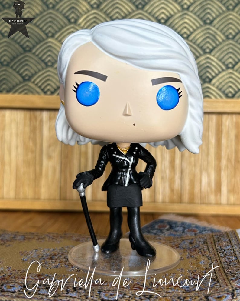 May include: A stylized vinyl collectible figure with white hair, bright blue eyes, and a small beauty mark. The figure wears a black leather-style jacket, a black skirt, and black boots, while holding a black cane with a silver handle.