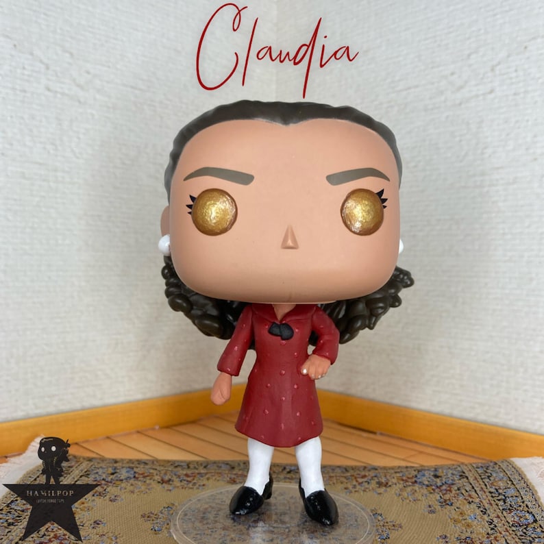 May include: A vinyl collectible figure named Claudia with dark brown curly hair, large golden eyes, and a small nose. The figure wears a long-sleeved dark red coat with a black collar detail over white leggings and black shoes, standing on a clear base.