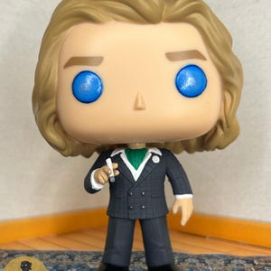 May include: A vinyl collectible figure featuring a character with wavy blonde hair and bright blue eyes. The figure wears a dark grey double-breasted suit with a white shirt, a green cravat, and a small white flower lapel pin, holding a small white cigarette.