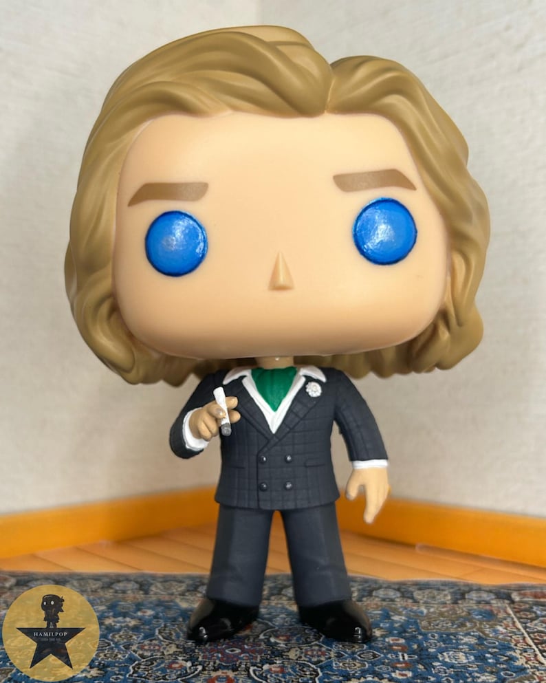 May include: A vinyl collectible figure featuring a character with wavy blonde hair and bright blue eyes. The figure wears a dark grey double-breasted suit with a white shirt, a green cravat, and a small white flower lapel pin, holding a small white cigarette.
