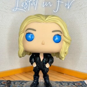May include: A vinyl collectible figure with blonde hair and bright blue eyes, wearing a black glittery coat over a black vest and pants. The figure stands on a clear circular base. The text "Lestat in Fur" is written in white script above the figure.