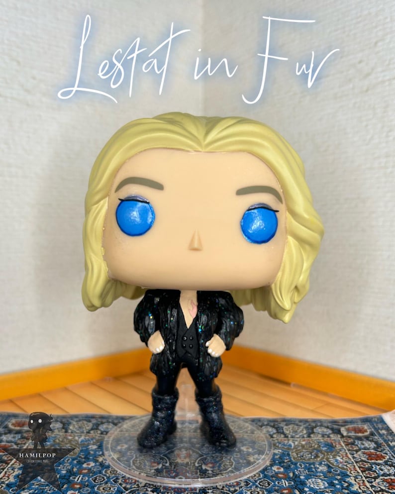 May include: A vinyl collectible figure with blonde hair and bright blue eyes, wearing a black glittery coat over a black vest and pants. The figure stands on a clear circular base. The text "Lestat in Fur" is written in white script above the figure.