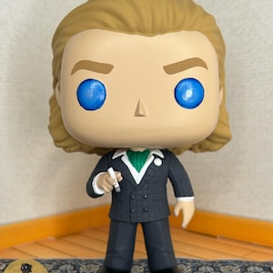 May include: A vinyl collectible figure featuring a character with light brown hair and bright blue eyes. The figure wears a dark gray double-breasted suit with a white shirt, a green cravat, and a white lapel flower, while holding a small white object.