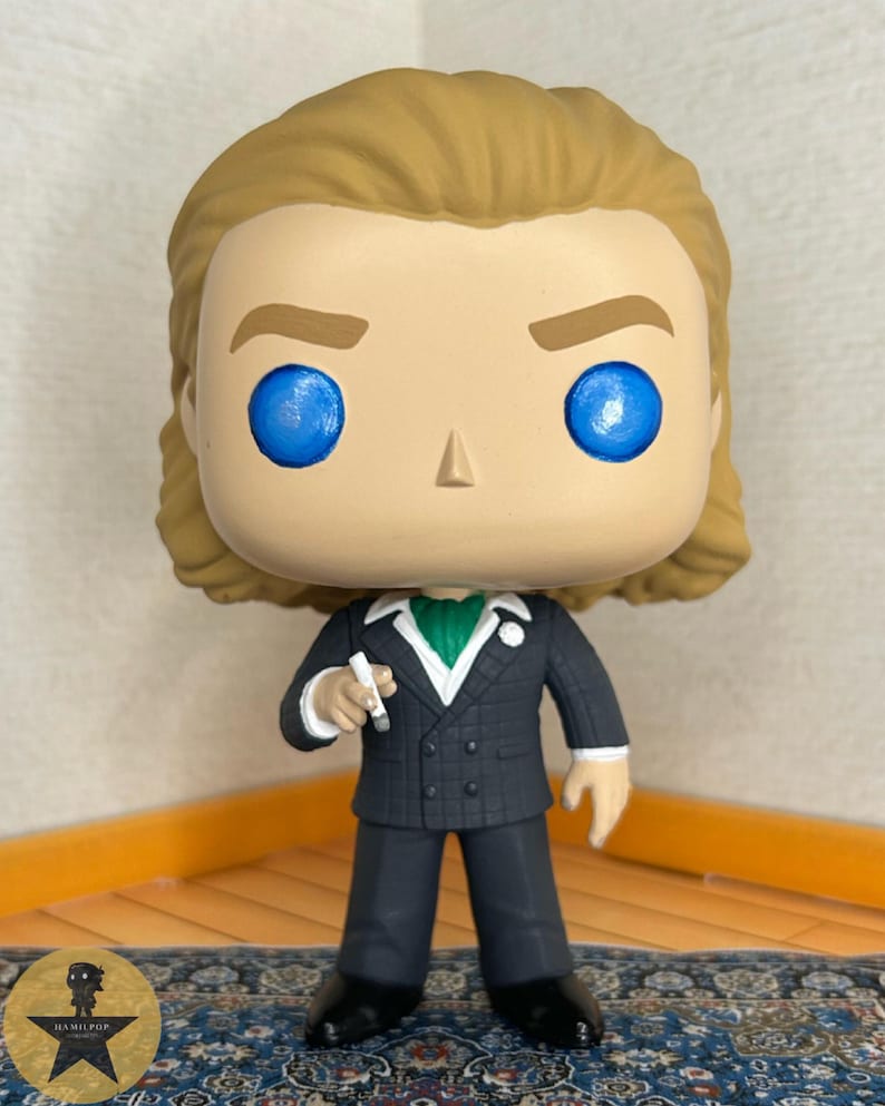 May include: A vinyl collectible figure featuring a character with light brown hair and bright blue eyes. The figure wears a dark gray double-breasted suit with a white shirt, a green cravat, and a white lapel flower, while holding a small white object.