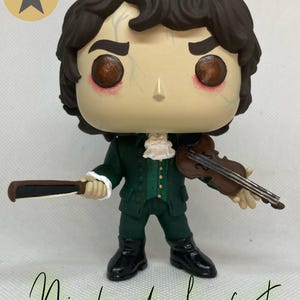 May include: A stylized vinyl figure of a character with dark brown hair, wearing a dark green coat with gold buttons, a white cravat, and black boots. The figure holds a brown violin and a bow. The text "Hamilpop" appears in a circle, and "Nicolas de Lenfent" is written below.