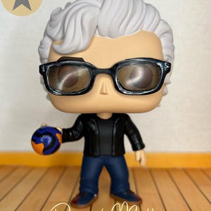 May include: A vinyl collectible figure featuring a character with white hair, black-rimmed glasses, a black jacket, and blue jeans. The figure holds a small, round, orange and blue patterned ball in one hand. The name Daniel Molloy is written below.