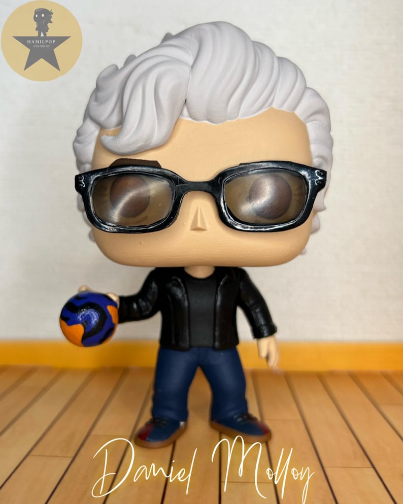 May include: A vinyl collectible figure featuring a character with white hair, black-rimmed glasses, a black jacket, and blue jeans. The figure holds a small, round, orange and blue patterned ball in one hand. The name Daniel Molloy is written below.