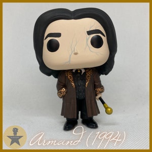 May include: A vinyl collectible figure featuring a character with black hair, pale skin with light blue vein details, wearing a brown coat with gold scroll patterns over a black vest and trousers, holding a black cane with a gold handle.