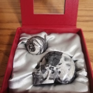 May include: Two small, intricately carved stone skulls in a red velvet box. One skull is a sphere, the other is a traditional skull shape. Both are white with black veins.