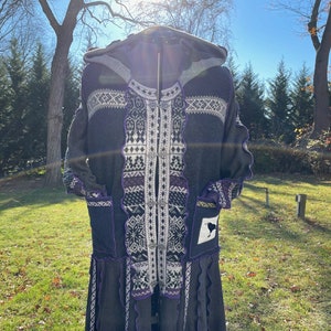 May include: A long, gray hooded cardigan with a white and purple geometric pattern. The cardigan has a ruffled hem and a black crow patch on the front.