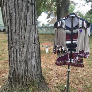 May include: A brown, beige, and burgundy patchwork jacket with a zipper closure and a hood. The jacket has a heart-shaped patch with purple glitter on the left side. The jacket is on a black stand against a backdrop of green grass and a tree.