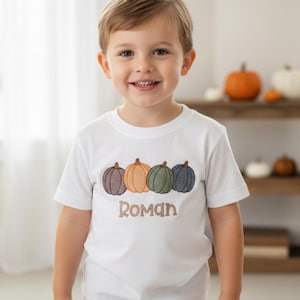 May include: White short-sleeve t-shirt with an embroidered design of four pumpkins in fall colors. The name "ROMAN" is embroidered below the pumpkins. The child is wearing blue jeans.