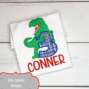 May include: White t-shirt featuring a green dinosaur holding a blue plaid number 3. The name "CONNER" is embroidered in red below the number. The shirt is a custom design.