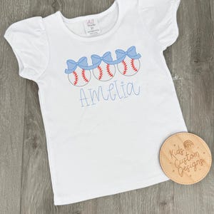 May include: White t-shirt with short, puffed sleeves. The shirt features an embroidered design of three baseballs with red stitching and blue bows. The name "Amelia" is embroidered in blue script below the baseballs.