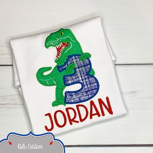 May include: White t-shirt featuring a green dinosaur with a blue plaid number 3. The name "JORDAN" is embroidered in red. The shirt is designed for a child's birthday. The image also includes the text "Kids Custom Designs".