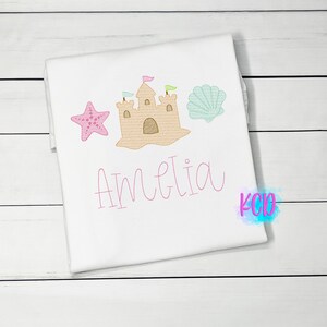 May include: White t-shirt with pink embroidery of a sandcastle, a starfish, and a seashell. The name Amelia is embroidered below the design.