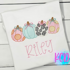 May include: A white t-shirt with a pink, orange, blue, and black and white pumpkin design. The pumpkins have bows and the text "Riley" is embroidered below the pumpkins.
