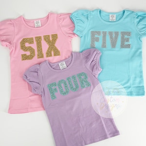 May include: Three girls' t-shirts with glitter numbers. The pink shirt says "SIX" in gold glitter, the blue shirt says "FIVE" in silver glitter, and the purple shirt says "FOUR" in teal glitter.
