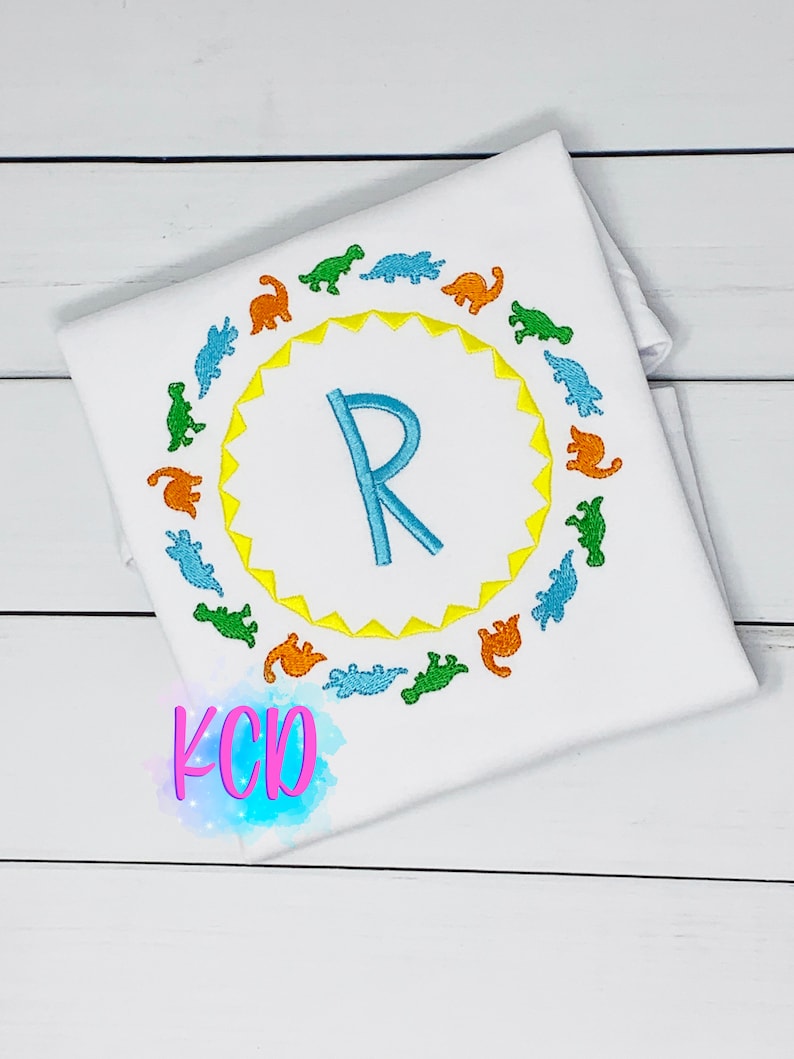 May include: White t-shirt with a blue letter R in a yellow sun with a circle of colorful dinosaurs around it. The text "KCD" is in pink on the bottom of the shirt.