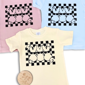 May include: Three pastel-coloured t-shirts with a black and white chequered pattern and a graphic of three cartoon Peeps wearing sunglasses. The text "Kids Custom Designs" is on a wooden tag.