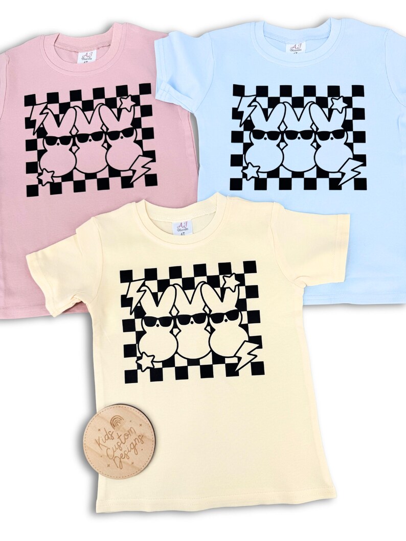 May include: Three pastel-coloured t-shirts with a black and white chequered pattern and a graphic of three cartoon Peeps wearing sunglasses. The text "Kids Custom Designs" is on a wooden tag.
