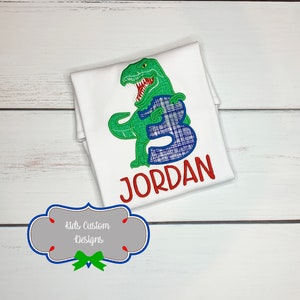 May include: White t-shirt with a green dinosaur design holding a blue plaid number 3. The name "JORDAN" is embroidered in red below the number. The shirt is on a white wooden surface.