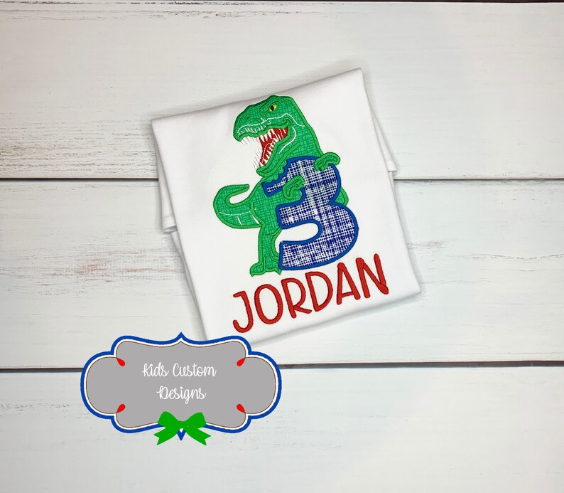 May include: White t-shirt with a green dinosaur design holding a blue plaid number 3. The name "JORDAN" is embroidered in red below the number. The shirt is on a white wooden surface.