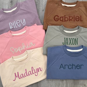 May include: A collection of six crewneck sweatshirts in various colors, each embroidered with a different name: Riley, Gabriel, Sophia, Jaxon, Madalyn, and Archer. The sweatshirts are in shades of lavender, brown, pink, gray, beige, and blue.