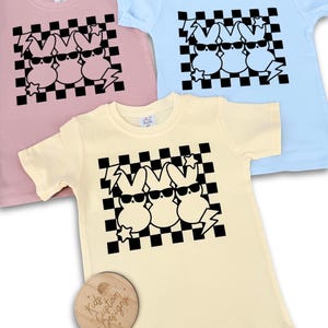 May include: Three black and white cartoon bunnies wearing sunglasses on a checkered background. The bunnies are on a yellow, pink, and light blue t-shirt. The t-shirts are for kids.