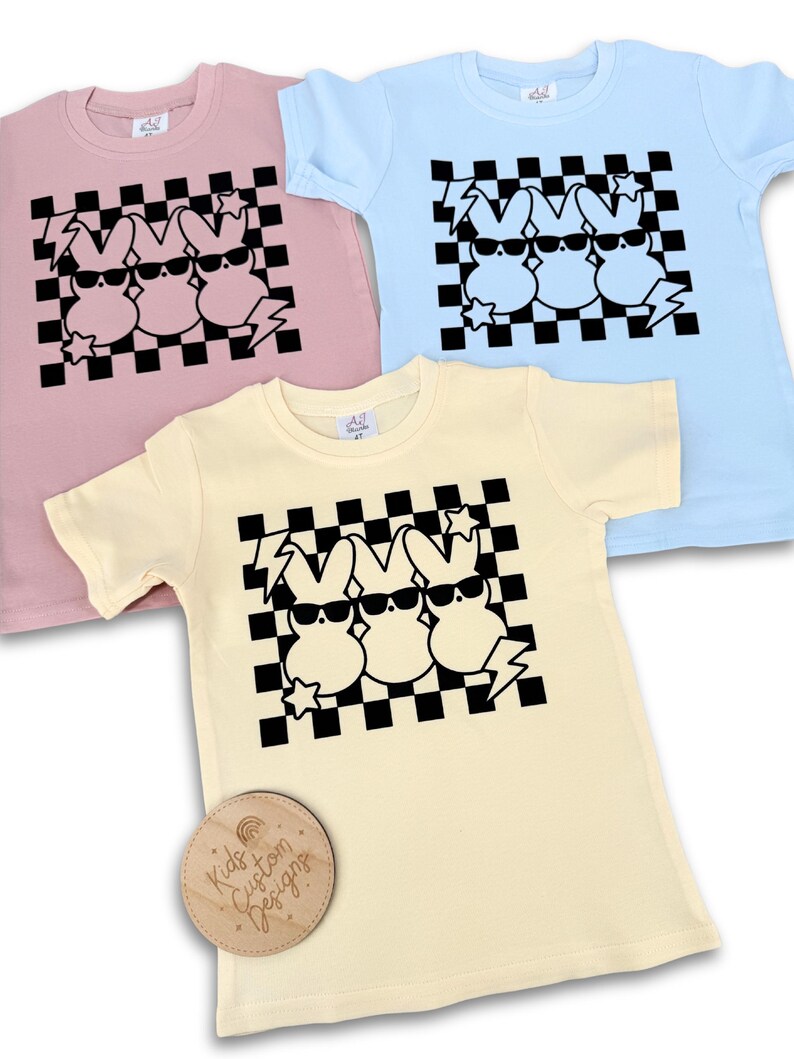 May include: Three black and white cartoon bunnies wearing sunglasses on a checkered background. The bunnies are on a yellow, pink, and light blue t-shirt. The t-shirts are for kids.