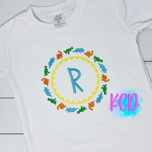 May include: White long-sleeve t-shirt with a blue letter R surrounded by a circle of colorful dinosaurs. The text "KCD" is visible in the bottom right corner of the image.