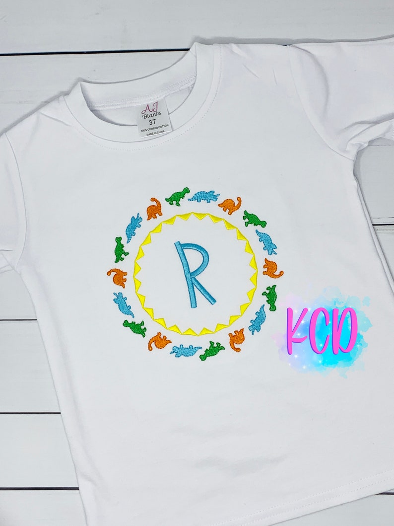 May include: White long-sleeve t-shirt with a blue letter R surrounded by a circle of colorful dinosaurs. The text "KCD" is visible in the bottom right corner of the image.