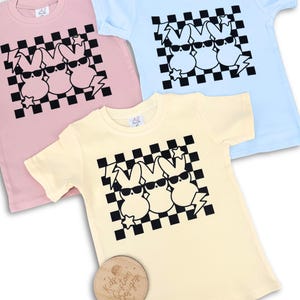 May include: Three pastel coloured t-shirts with a black and white chequered pattern and a graphic of three cartoon bunnies wearing sunglasses. The bunnies are in a row and have a lightning bolt and star on either side of them. The t-shirts are pink, light blue, and yellow.