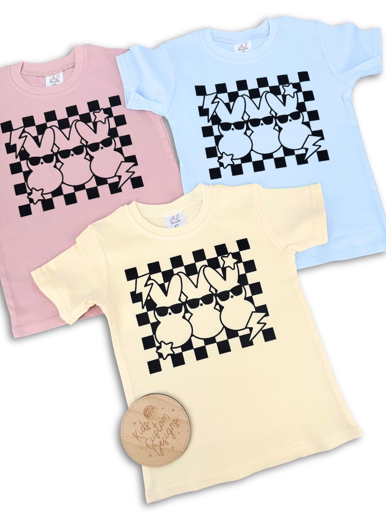 May include: Three pastel coloured t-shirts with a black and white chequered pattern and a graphic of three cartoon bunnies wearing sunglasses. The bunnies are in a row and have a lightning bolt and star on either side of them. The t-shirts are pink, light blue, and yellow.