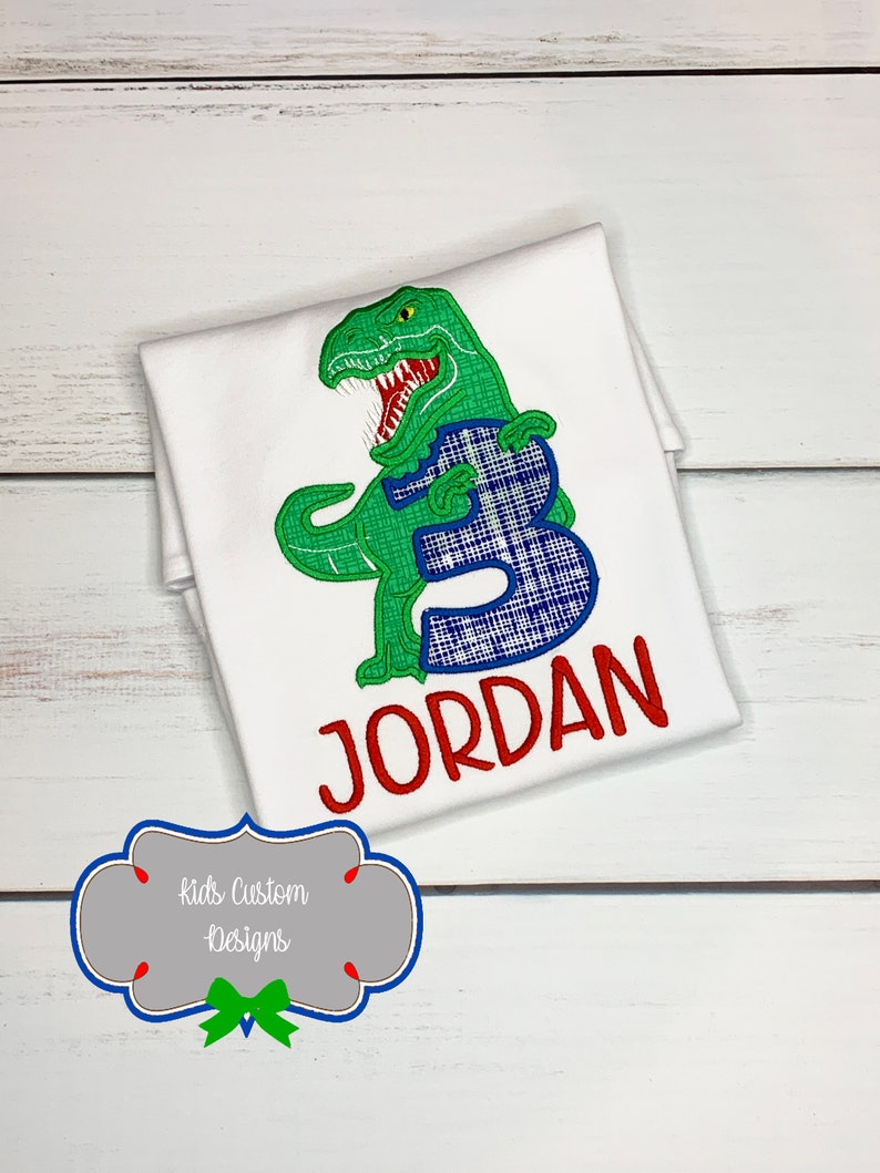 May include: White t-shirt featuring a green dinosaur with a blue plaid number 3. The name "JORDAN" is embroidered in red. The shirt is designed for a child's birthday. The image also includes the text "Kids Custom Designs".