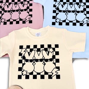 May include: Three pastel coloured t-shirts with a black and white chequered pattern and three cartoon bunnies wearing sunglasses. The bunnies are wearing sunglasses and have a lightning bolt and star design.