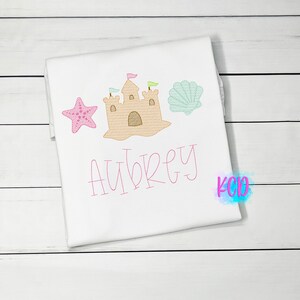 May include: White t-shirt with pink embroidery of a sandcastle, a starfish, and a seashell. The name "Aubrey" is embroidered below the sandcastle.